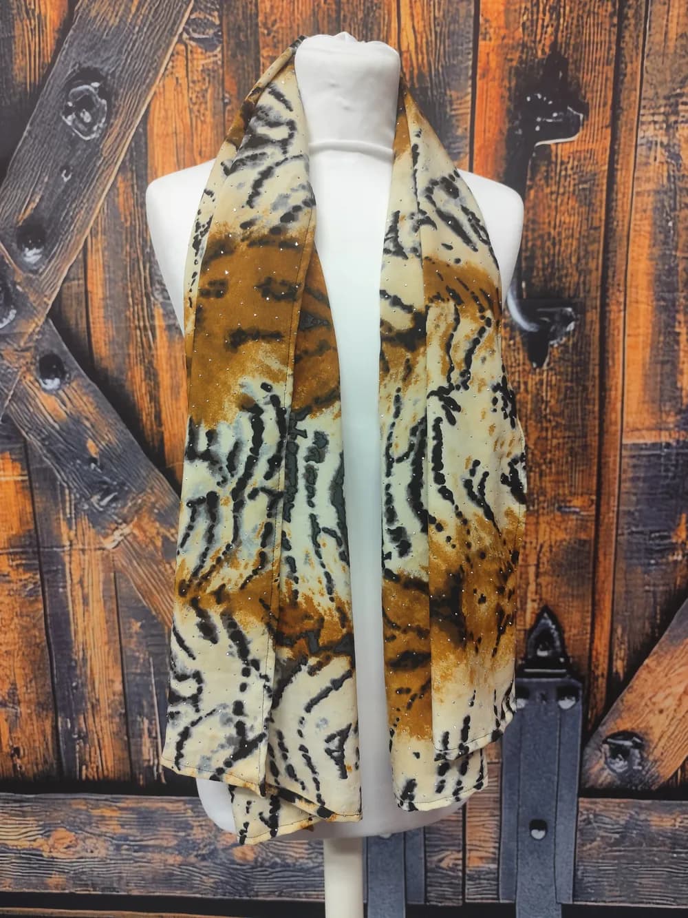 Foulard à motif tigré à dominante brun roux, rayures noires et fond crème, semé de minuscules strass argentés concentrés sur les bandes brun roux, noué simplement autour du cou d'un buste mannequin, deux pans longs retombant devant une porte en bois orangée