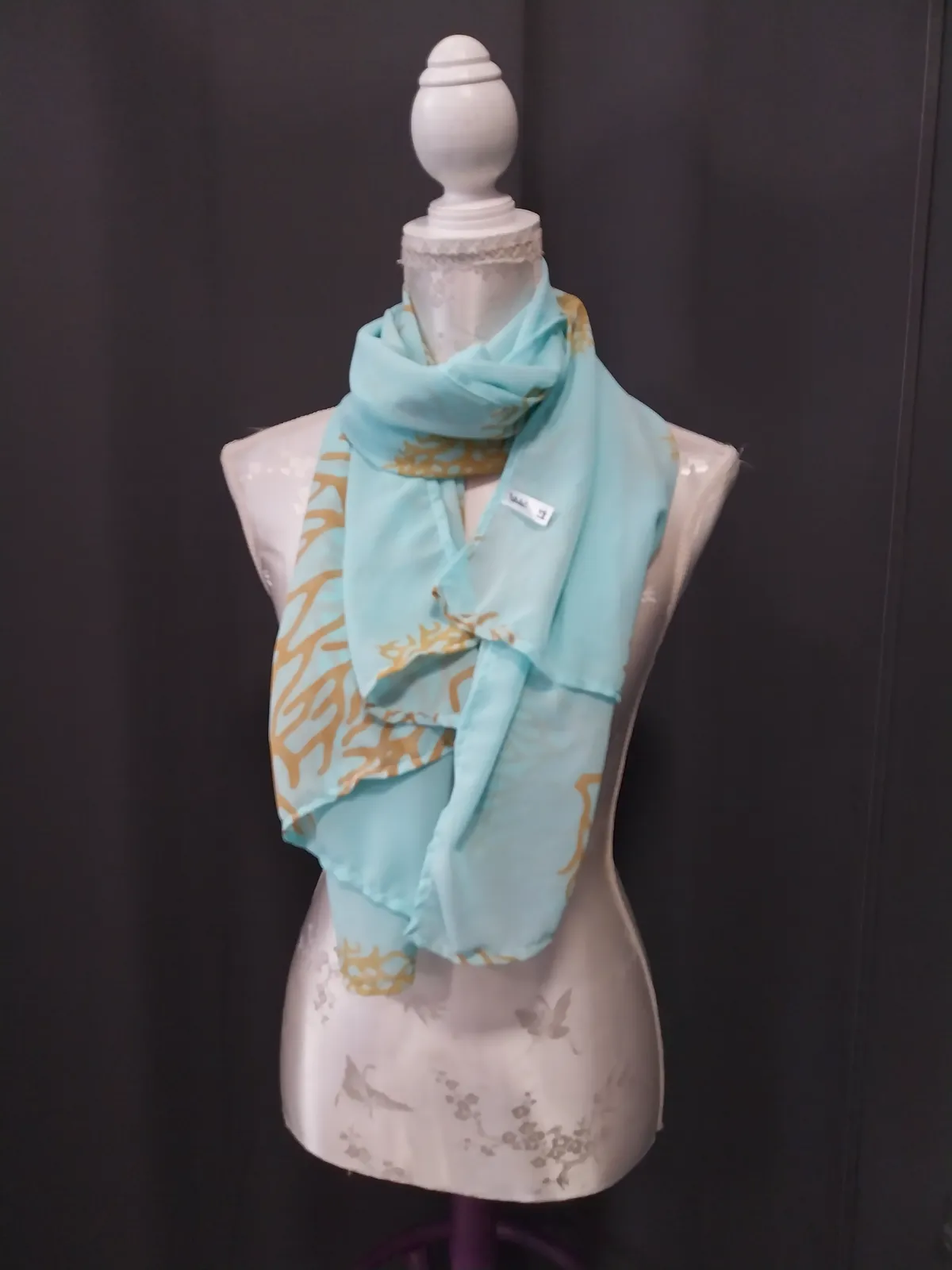 Foulard vert d'eau à motif corail doré, noué autour du cou d'un buste mannequin, étiquette tissée visible sur le pan gauche, fond sombre