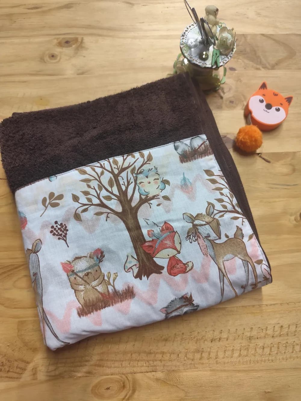 Linge en éponge marron chocolat, plié et posé en biais sur une table en bois clair. Le haut montre l'éponge bouclée d'un brun profond. Le bas est couvert d'une bande de coton blanc cousue, imprimée d'animaux de la forêt aux couleurs douces : au centre, un arbre aux feuilles brunes avec une petite chouette bleue perchée dans ses branches, un renard roux assis à son pied, un ourson brun à gauche et une biche à droite, plusieurs portant un bandeau à plumes. Des zigzags roses et gris pâle, des feuilles et des plumes complètent le décor. Aucun prénom n'est brodé. Derrière, un pot à ciseaux décoré et un mètre ruban en forme de renard orange.