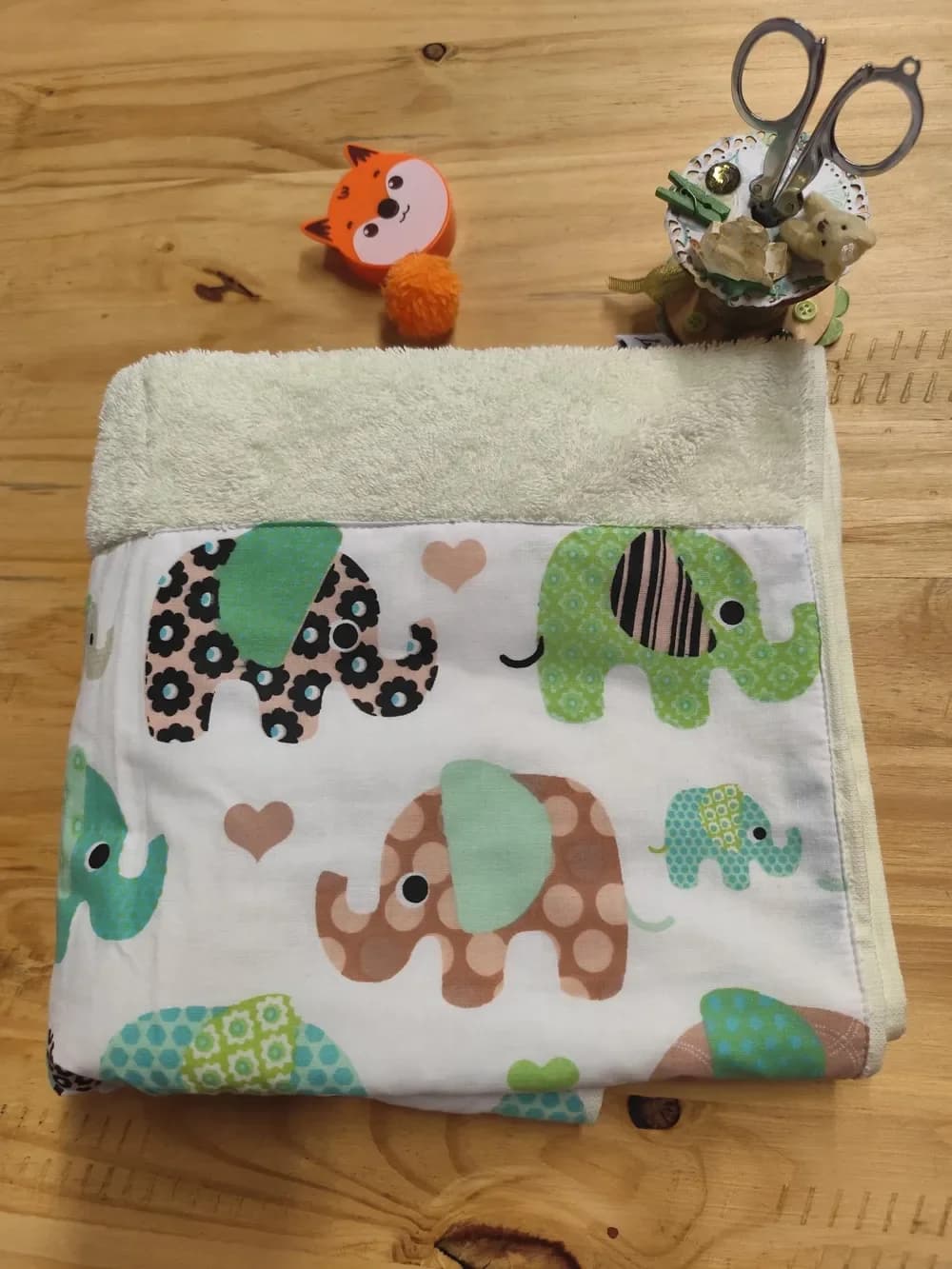 Linge en éponge pour bébé fait main CÉKIKAFÉ, plié et posé à plat sur une table en bois, vu de face : en haut l'éponge épaisse vert d'eau très pâle, et sur le bas une large bande de coton blanc imprimée d'éléphants multicolores, à fleurs et marron : un éléphant vert à oreille rayée, un éléphant noir à fleurs sur fond rose, un éléphant marron à pois, un éléphanteau turquoise, entre de petits cœurs roses. Aucun prénom n'est encore brodé.