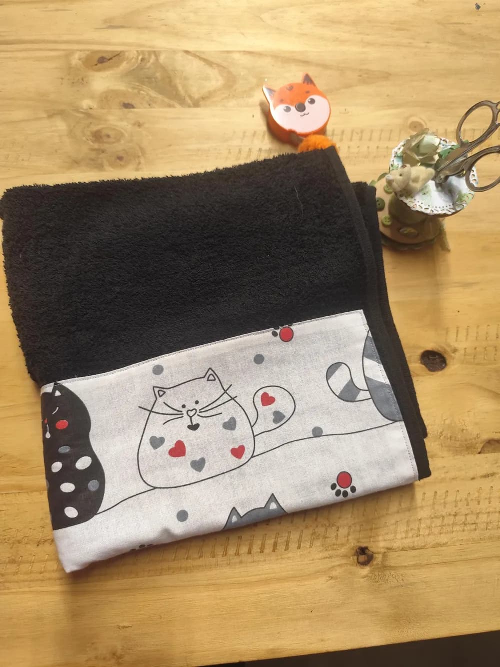 Linge en éponge noire, plié et posé sur une table en bois clair. Le haut montre l'éponge bouclée d'un noir profond. Le bas est couvert d'une bande de coton blanc cousue, imprimée de chats dessinés façon bande dessinée : au centre, un gros chat blanc au trait noir, couché sur une ligne, le ventre décoré de cœurs rouges et gris. Autour, un chat noir à pois blancs, une queue rayée grise, des pois gris et des empreintes de pattes rouges. Aucun prénom n'est brodé. Derrière le linge, un mètre ruban en forme de renard orange et un petit pot à ciseaux décoré.