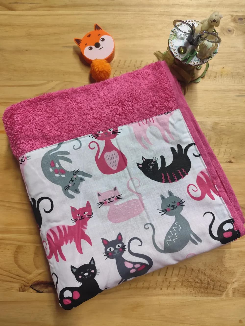 Linge en éponge rose vif, presque fuchsia, plié et posé en biais sur une table en bois clair. Le haut montre l'éponge bouclée rose vif. Le bas est couvert d'une large bande de coton blanc cousue, imprimée de nombreux chats dessinés : des chats roses, fuchsia, gris et noirs, certains assis la queue enroulée, d'autres qui marchent, d'autres encore couchés sur le dos ou endormis, les yeux fermés et les joues roses. Aucun prénom n'est brodé. Derrière le linge, un mètre ruban en forme de renard orange et un pot à ciseaux décoré.