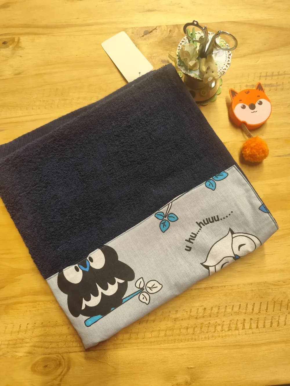 Linge en éponge bleu marine, plié et posé en biais sur une table en bois clair, avec une étiquette blanche qui dépasse en haut. Le haut montre l'éponge bouclée d'un bleu marine profond. Le bas est couvert d'une bande de coton gris-bleu clair cousue, imprimée de chouettes : à gauche, une chouette noire aux grands yeux perchée sur une branche turquoise ; à droite, une chouette blanche qui ferme les yeux, avec les mots « u hu… huuu ». Des petites feuilles turquoise sont semées autour. Aucun prénom n'est brodé. À côté, un pot à ciseaux décoré et un mètre ruban en forme de renard orange.