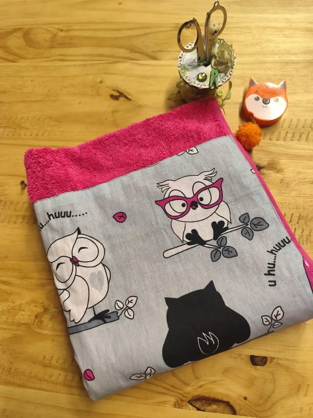 Linge en éponge rose vif, plié et posé en biais sur une table en bois clair. Le haut montre l'éponge bouclée rose vif. Le bas est couvert d'une large bande de coton gris clair cousue, imprimée de chouettes blanches et noires perchées sur des branches feuillues : une chouette blanche porte de grandes lunettes rose vif, une autre ferme les yeux d'un air rêveur, une troisième, toute noire, est vue de dos. Les mots « u hu… huuu » sont écrits entre elles. Aucun prénom n'est brodé. À côté, un mètre ruban en forme de renard orange et un petit pot à ciseaux décoré.