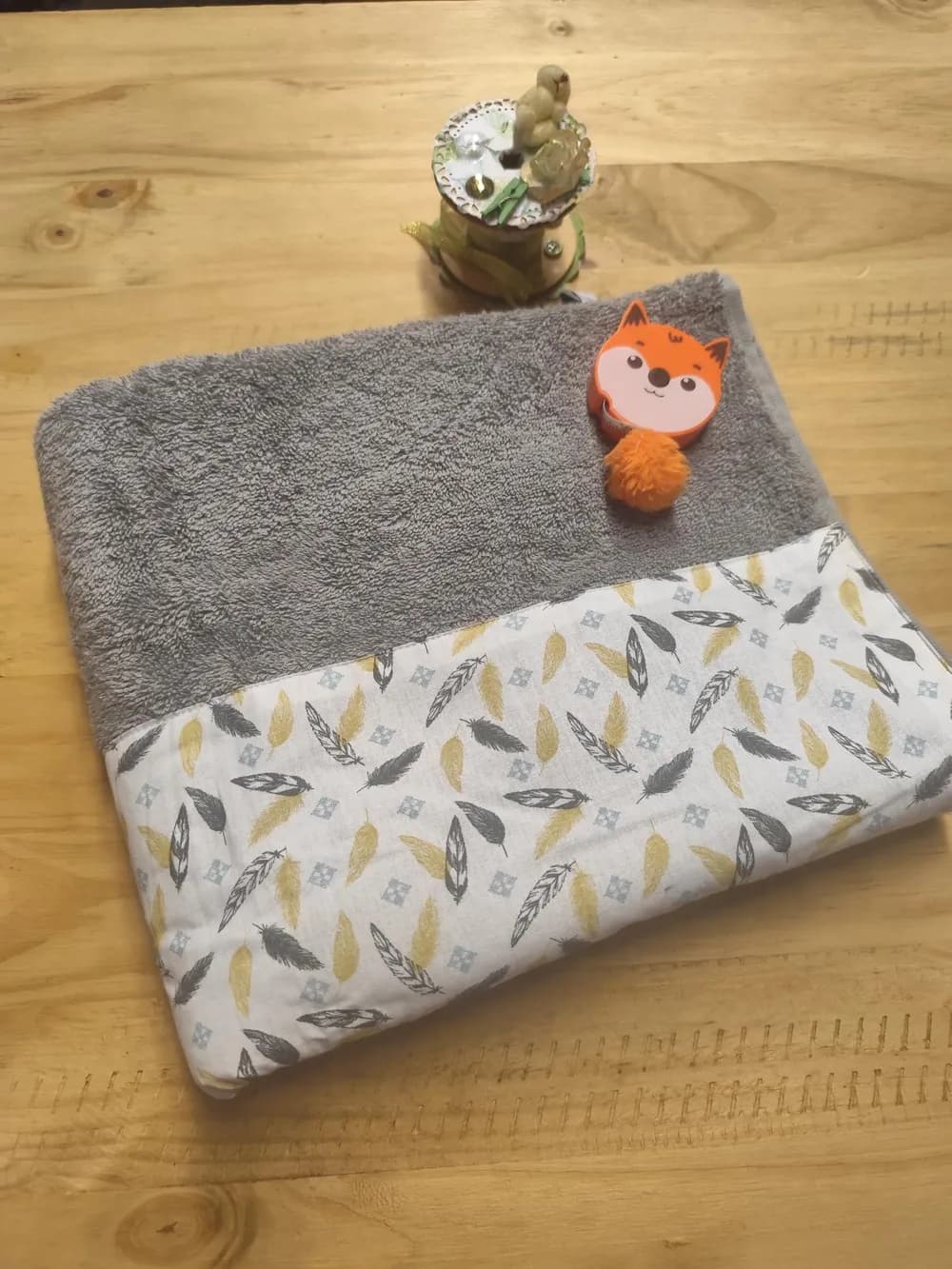 Linge en éponge gris moyen, plié et posé en biais sur une table en bois clair. Le haut montre l'éponge bouclée d'un gris doux et uni. Le bas est couvert d'une bande de coton blanc cousue, imprimée de nombreuses plumes éparpillées en tous sens : des plumes dorées et ocre, des plumes gris foncé presque noires, et des plumes blanches au contour noir finement strié, parsemées de petits carrés gris-bleu. Aucun prénom n'est brodé. Un mètre ruban en forme de renard orange est posé sur l'éponge, et un pot à ciseaux décoré se trouve derrière le linge.