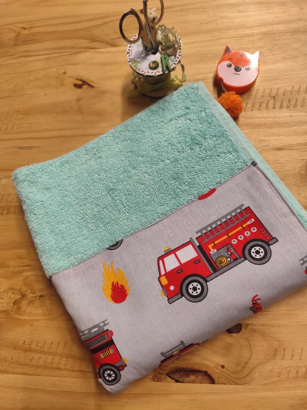 Linge en éponge vert d'eau, plié et posé en biais sur une table en bois clair. Le haut montre l'éponge bouclée, d'un vert d'eau doux tirant sur le menthe. Le bas est couvert d'une bande de coton gris clair cousue, imprimée de camions de pompiers rouges : au centre, un grand camion rouge vu de profil, avec son échelle grise et son tuyau enroulé jaune, à côté d'une flamme rouge et jaune. D'autres camions dépassent sur les bords. Aucun prénom n'est brodé. Derrière le linge, un pot à ciseaux décoré et un mètre ruban en forme de renard orange.