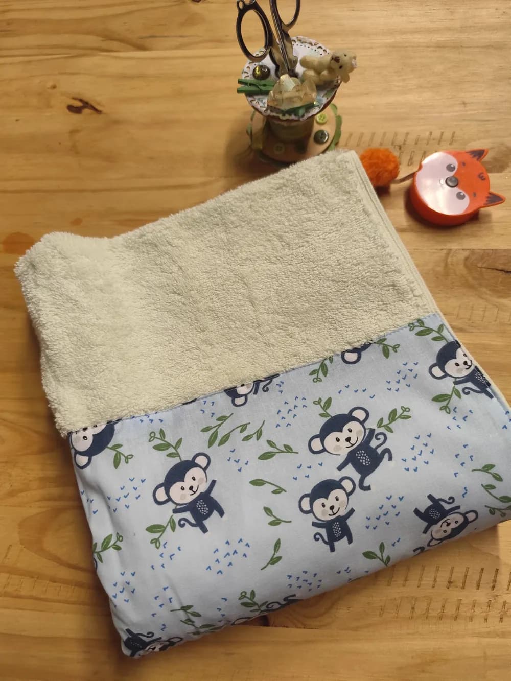 Linge en éponge vert anis très clair, presque crème, plié et posé en biais sur une table en bois. Le haut montre l'éponge bouclée d'un vert tendre et pâle. Le bas est couvert d'une bande de coton bleu ciel cousue, imprimée de petits singes bleu gris foncé au visage clair et souriant, qui dansent, se balancent ou font la roue, entourés de brindilles aux feuilles vertes et de petits traits bleus. Aucun prénom n'est brodé. Derrière le linge, un pot à ciseaux décoré et un mètre ruban en forme de renard orange.