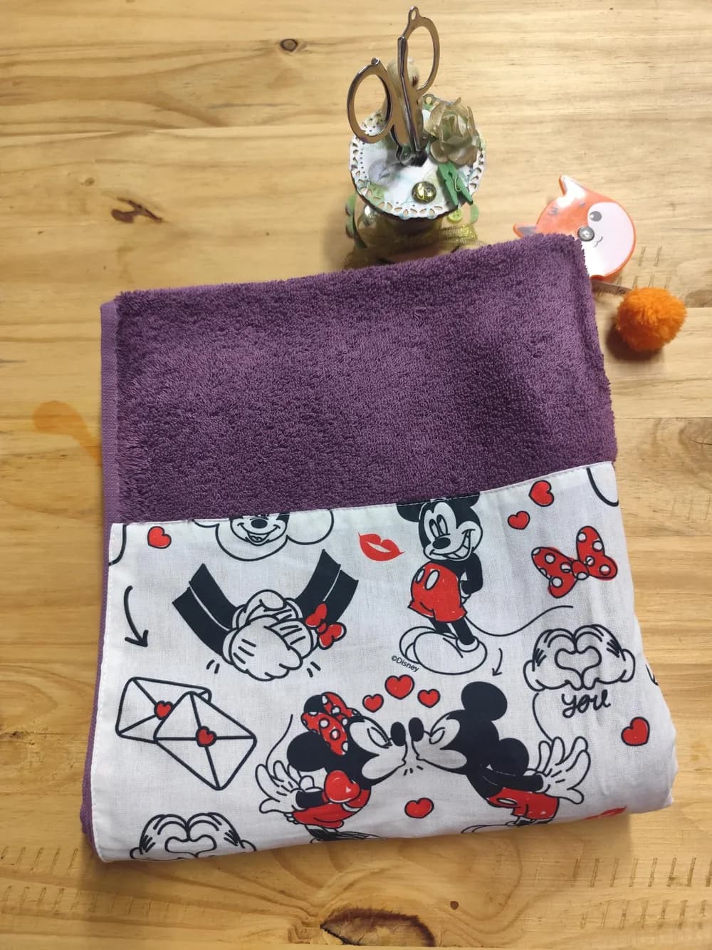 Linge en éponge violet prune, plié et posé sur une table en bois clair. Le haut montre l'éponge bouclée d'un violet profond et chaleureux. Le bas est couvert d'une bande de coton blanc cousue, imprimée en noir et rouge de petites souris de dessin animé aux grandes oreilles rondes : l'une en short rouge sourit, deux autres s'embrassent entourées de petits cœurs rouges. Autour, des nœuds papillon rouges à pois blancs, des bouches rouges, des cœurs, des enveloppes scellées d'un cœur et des mains formant un cœur avec le mot « you ». Aucun prénom n'est brodé. Derrière, un pot à ciseaux décoré et un mètre ruban en forme de renard orange.