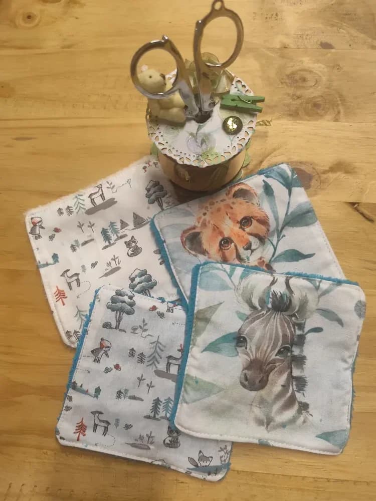 Lingettes lavables bébé motifs animaux, 10 x 10 cm, éponge de bambou – CÉKIKAFÉ. Choix 1, côté imprimé : un bébé guépard et un zèbre à l'aquarelle parmi des feuilles vertes, et deux lingettes blanches à petits motifs de forêt, de faons et de renards, bordées d'éponge bleue, sur une table en bois sous une bobine de couture et des ciseaux.