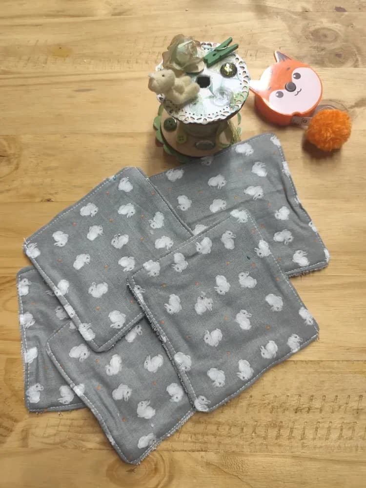 Lingettes lavables bébé en coton gris à lapins blancs, éponge de bambou grise – CÉKIKAFÉ. Cinq lingettes étalées côté imprimé sur une table en bois : coton gris semé de petits lapins blancs et de minuscules points orange. En haut, une bobine de couture décorée d'un petit ourson et un mètre ruban orange en forme de tête de renard avec son pompon.