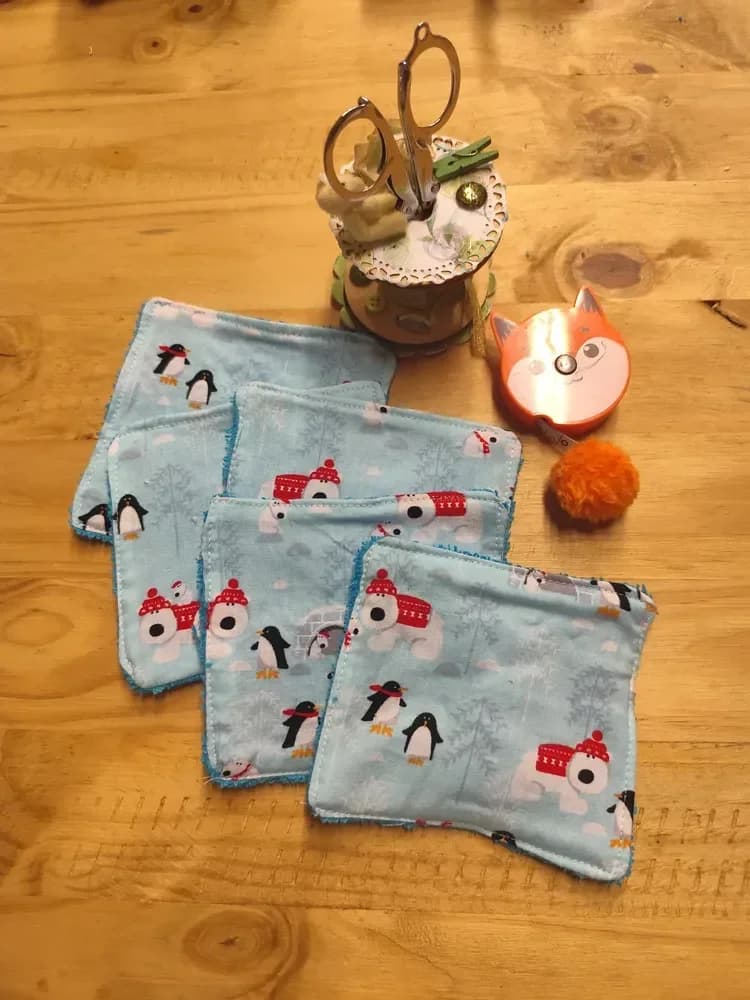 Lingettes lavables bébé pingouins et ours polaires, bonnet et pull rouges, flocons de neige, éponge turquoise – CÉKIKAFÉ. Cinq lingettes étalées côté imprimé sur une table en bois : coton bleu ciel orné de petits pingouins à écharpe rouge, d'ours polaires blancs en bonnet rouge à pompon et pull rouge, d'un igloo, de sapins enneigés et de flocons, avec une bordure d'éponge turquoise. En haut, une bobine de couture décorée avec des ciseaux et un mètre ruban orange en forme de tête de renard.
