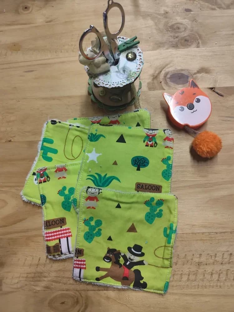 Cinq lingettes lavables carrées étalées en éventail sur une table en bois, côté coton jaune vert vif. L'imprimé Far West montre un petit cow-boy à chapeau haut-de-forme sur un cheval brun, des panneaux « SALOON », des façades rayées rouge et blanc, des cactus verts, des étoiles blanches, des lassos et de petits personnages. En arrière-plan, une bobine de couture décorée avec des ciseaux argentés et un petit ourson, et un mètre ruban orange en forme de tête de renard avec son pompon orange.