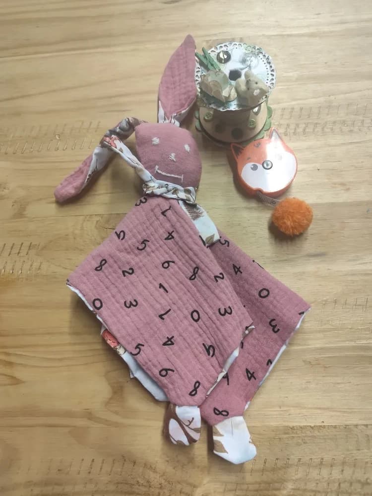 Mon doudou lapin rose CÉKIKAFÉ posé sur une table en bois clair, côté gaze de coton vieux rose imprimée de chiffres noirs. En haut, la tête ronde en gaze rose aux yeux et à la bouche brodés en fil blanc, avec deux grandes oreilles, l'une dressée, l'autre couchée, roses d'un côté et en coton blanc imprimé de l'autre, et une petite écharpe en coton imprimé nouée au cou. Le carré est replié et laisse voir des pattes en coton blanc imprimé aux coins. À côté, une bobine décorée et un mètre ruban en forme de renard orange.