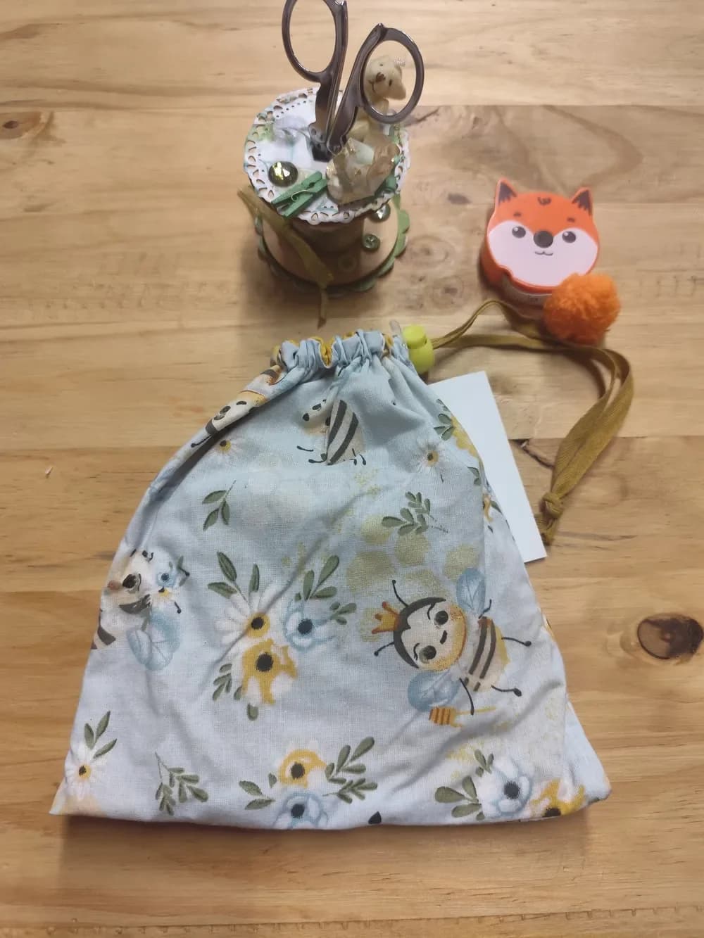 Pochon fait main CÉKIKAFÉ en coton bleu ciel très pâle, imprimé de petites abeilles rayées jaune et noir, de marguerites et de fleurs jaunes et bleues, fermé par un cordon en coton ocre et un serre-cordon en plastique jaune. On aperçoit la doublure jaune au niveau des fronces. Posé sur une table en bois.