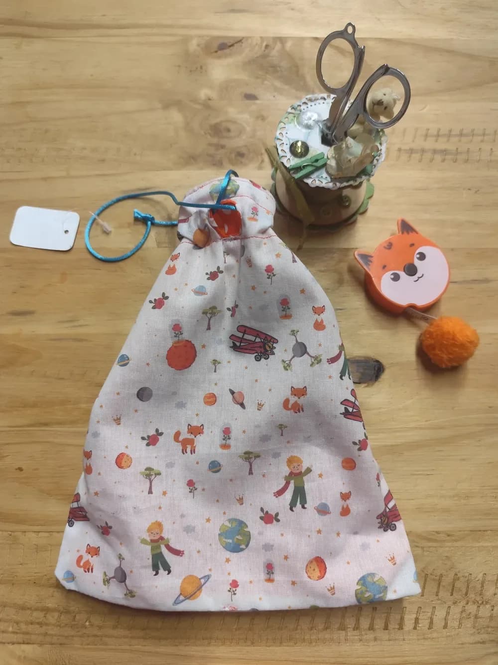 Pochon fait main CÉKIKAFÉ fermé et froncé en haut, posé sur une table en bois. Son coton blanc rosé est imprimé de l'univers du Petit Prince : le petit garçon aux cheveux blonds et à l'écharpe rouge, des renards orange, des avions rouges, des roses sous cloche, des planètes et des baobabs. Le cordon bleu turquoise passe dans un petit serre-cordon en tissu orange sur le devant.