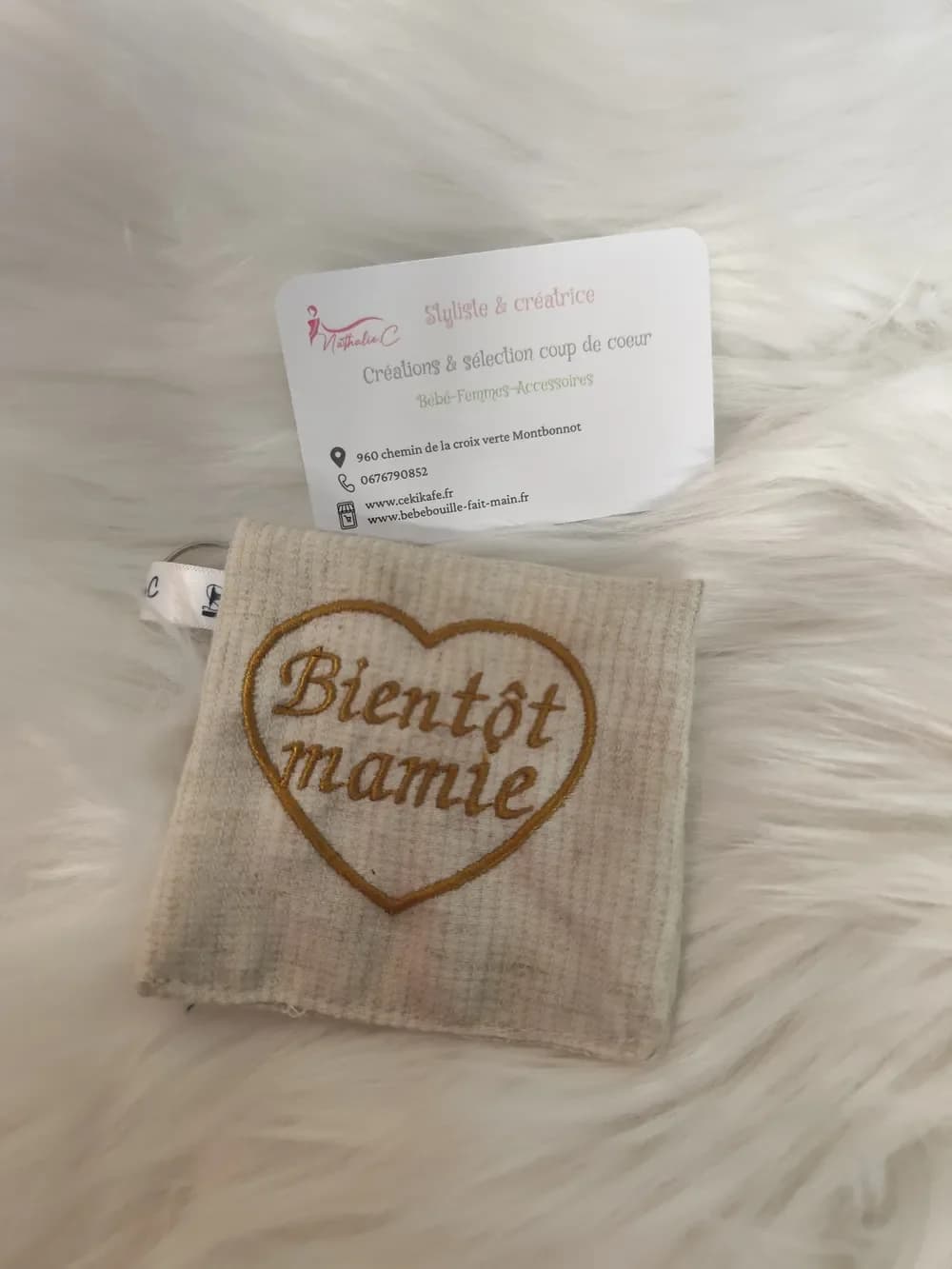 Porte-clé fait main CÉKIKAFÉ en coton belge, brodé d'un cœur et de l'inscription « Bientôt mamie » : une petite pochette presque carrée en tissu gaufré beige naturel, avec un grand cœur et les mots « Bientôt mamie » en lettres cursives brodés en fil doré moutarde. En haut à gauche, un ruban de satin blanc marqué au nom de la créatrice retient l'anneau porte-clés. Le porte-clé est posé sur une fausse fourrure blanche, devant la carte de visite de Nathalie C, styliste et créatrice.