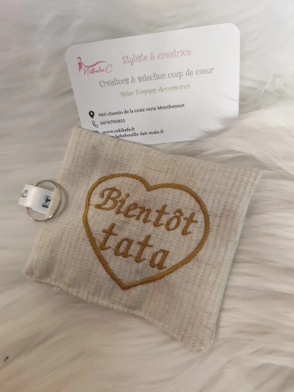 Porte-clé fait main CÉKIKAFÉ en coton, brodé d'un cœur et de l'inscription « Bientôt tata » : une petite pochette rectangulaire en tissu gaufré beige naturel, avec un grand cœur et les mots « Bientôt tata » en lettres cursives brodés en fil doré moutarde. Sur le côté gauche, un ruban de satin blanc marqué au nom de la créatrice retient un anneau porte-clés argenté. Le porte-clé est posé sur une fausse fourrure blanche, devant la carte de visite de Nathalie C, styliste et créatrice.