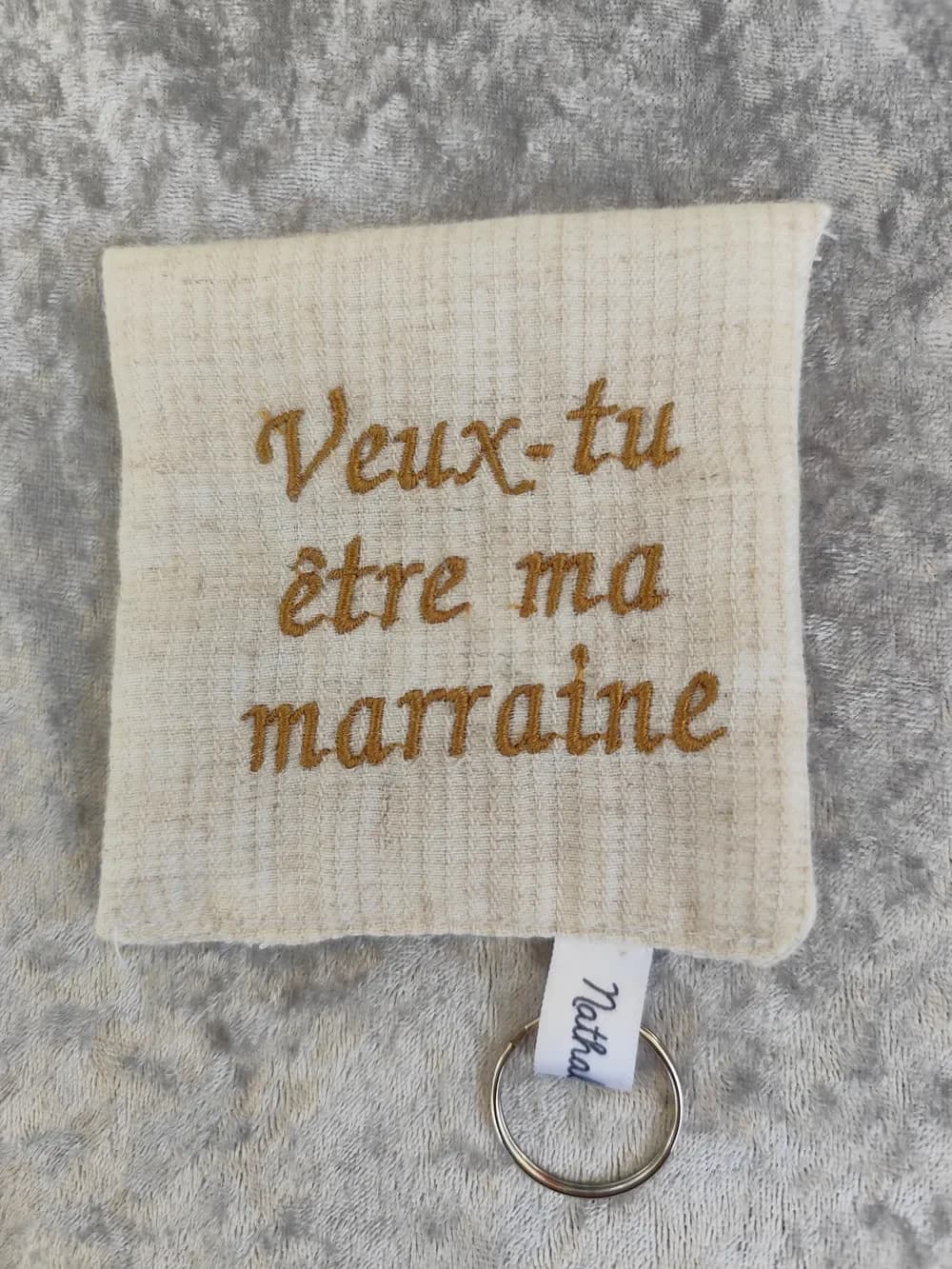 Porte-clé fait main CÉKIKAFÉ en coton belge, vu de face, à plat sur un velours gris argenté : une petite pochette presque carrée en coton gaufré beige naturel, brodée en fil doré moutarde, en lettres cursives sur trois lignes, de l'inscription « Veux-tu être ma marraine », sans cœur. En bas, un ruban de satin blanc marqué « Nathalie C » retient un anneau porte-clés argenté.