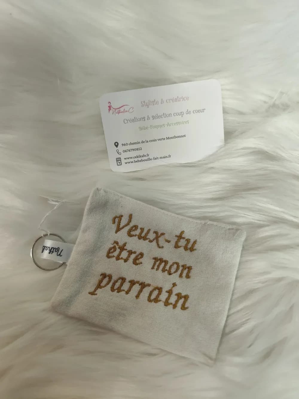 Porte-clé fait main CÉKIKAFÉ en coton belge, vu de face : une petite pochette rectangulaire en toile de coton écrue, brodée en fil doré moutarde, en lettres cursives sur trois lignes, de l'inscription « Veux-tu être mon parrain », sans cœur. Sur le côté gauche, un ruban de satin blanc marqué « Nathalie C » retient un anneau porte-clés argenté. Le porte-clé est posé sur une fausse fourrure blanche, sous la carte de visite de Nathalie C, styliste et créatrice.