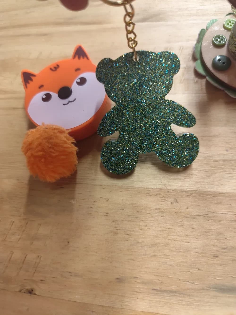 Porte-clés CÉKIKAFÉ en forme d'ourson en résine époxy pailletée, posé debout sur une table en bois clair : il est couvert de fines paillettes vertes, dorées et turquoise, sans dégradé, avec un effet brillant scintillant. En haut, une petite chaîne dorée part de l'oreille vers l'anneau porte-clés doré. À gauche, un mètre ruban orange en forme de tête de renard avec un pompon orange ; à droite, le bord d'une bobine décorée de boutons verts.