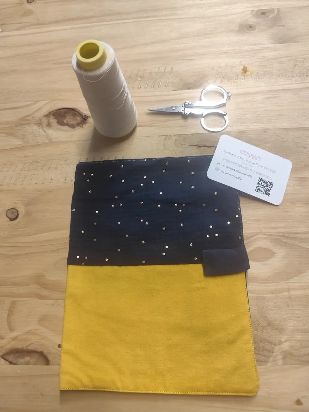 Protège-carnet de santé fermé, partie supérieure en tissu bleu marine semé de petits points pailletés dorés façon ciel étoilé, partie inférieure en tissu jaune moutarde uni, bande de fermeture scratch visible à la jonction des deux tissus, posé à plat sur une table en bois à côté d'une bobine de fil blanc, d'une paire de ciseaux et de la carte de visite CÉKIKAFÉ
