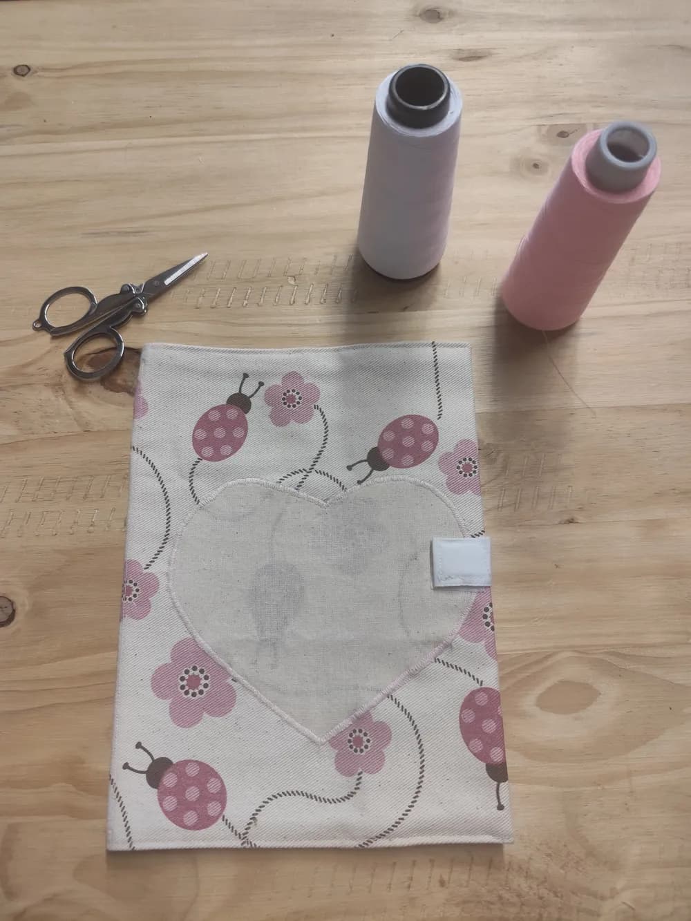 Protège-carnet de santé fermé posé à plat sur une table en bois, tissu écru imprimé de coccinelles roses à pois et de petites fleurs roses reliées par des pointillés, grand cœur appliqué au centre, languette de fermeture scratch blanche sur le bord droit, une bobine de fil blanc et une bobine de fil rose ainsi qu'une paire de ciseaux posées au-dessus