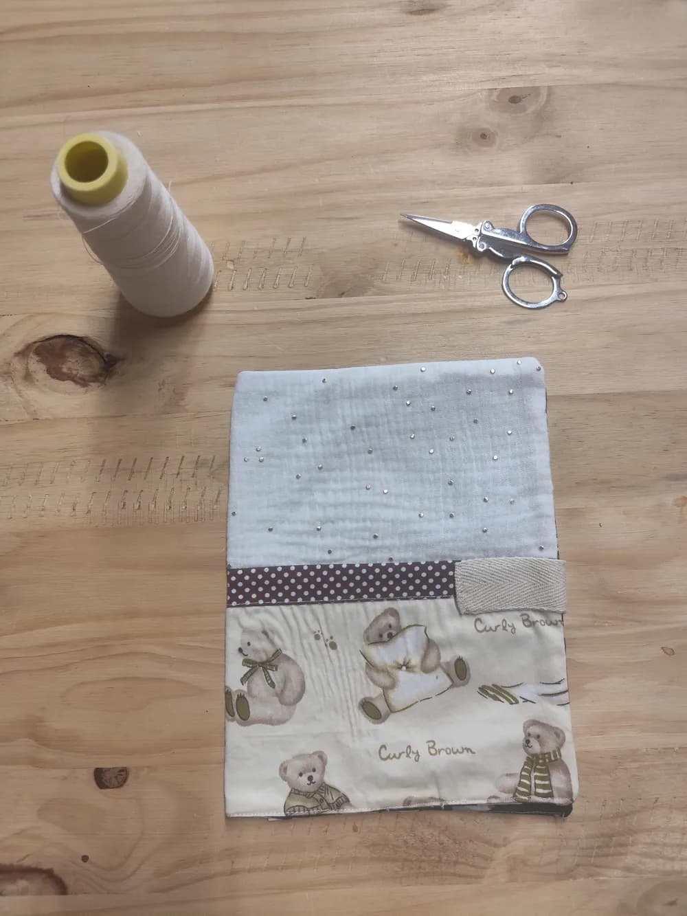 Protège-carnet de santé fermé posé à plat sur une table en bois, partie supérieure en gaze de coton blanche semée de petits strass brillants, biais marron à petits pois blancs au centre avec une languette de fermeture scratch beige, partie inférieure en coton imprimé de petits oursons « Curly Brown » écharpés, une bobine de fil blanc et une paire de ciseaux posées à côté