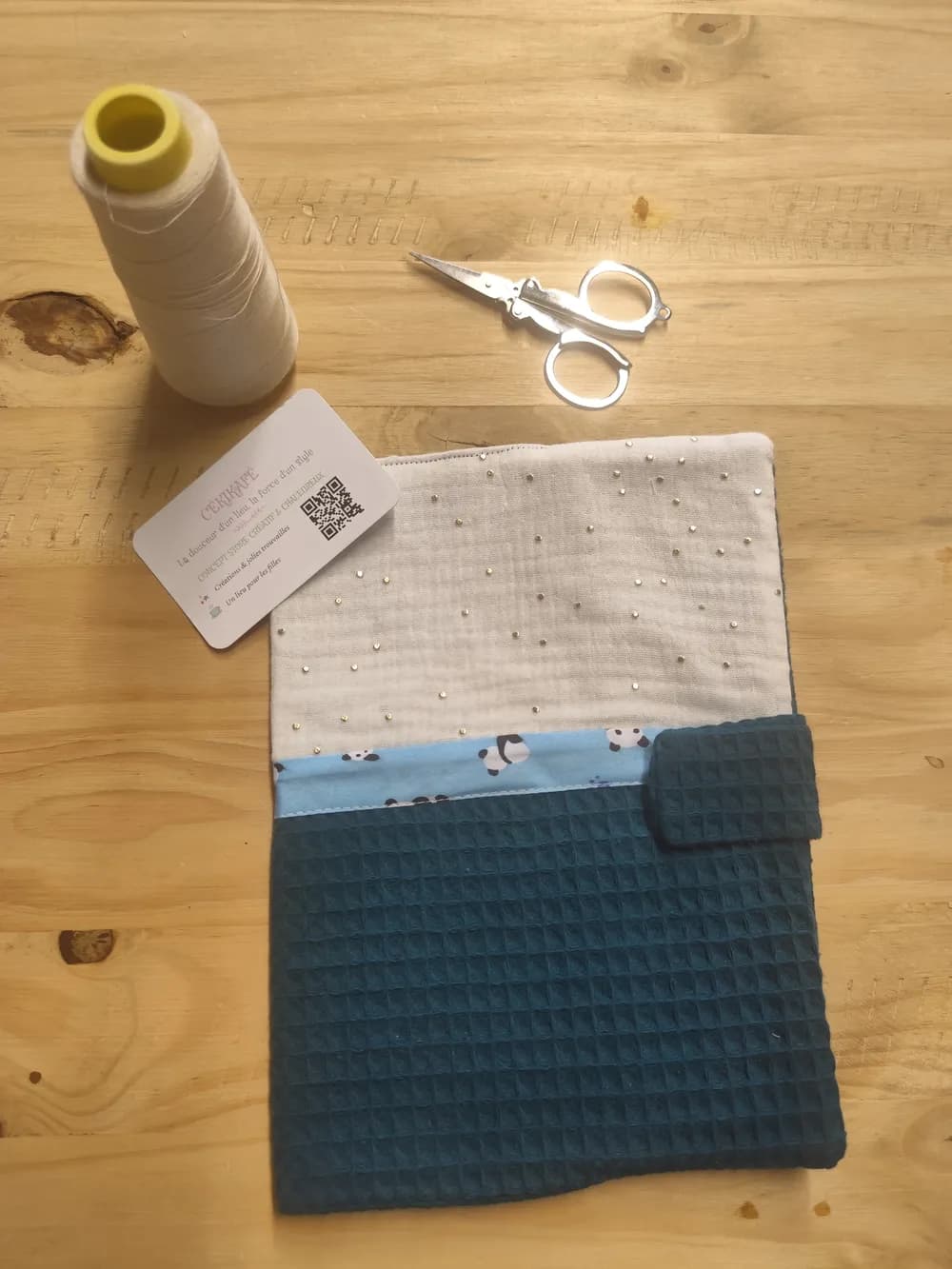 Protège-carnet de santé fermé posé à plat sur une table en bois, partie supérieure en gaze de coton blanche semée de petits strass brillants, biais bleu clair à petits pandas au centre, partie inférieure en coton texturé nid d'abeille bleu canard avec languette de fermeture scratch assortie sur le bord droit, une bobine de fil blanc, une paire de ciseaux et la carte de visite CÉKIKAFÉ posées au-dessus