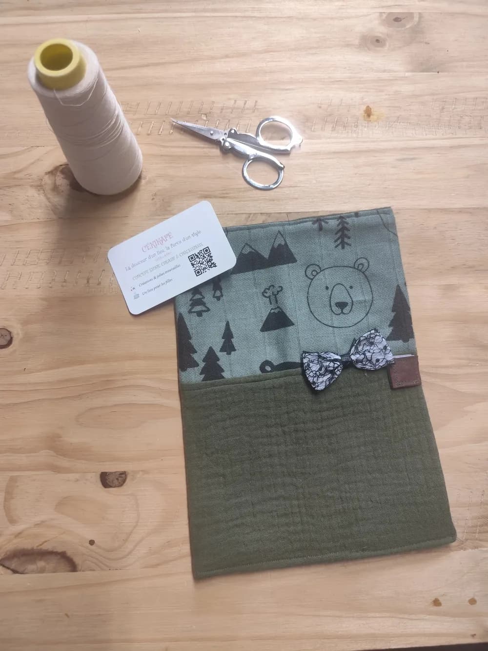 Protège-carnet de santé fermé posé à plat sur une table en bois, partie supérieure en gaze de coton kaki clair illustrée au trait noir d'une tête de nounours, de sapins et d'une montagne, partie inférieure en gaze de coton kaki uni, petit nœud cousu à motif blanc et noir sur le devant à la jonction des deux tissus, languette de fermeture scratch marron sur le bord droit, une bobine de fil blanc, une paire de ciseaux et la carte de visite CÉKIKAFÉ posées au-dessus