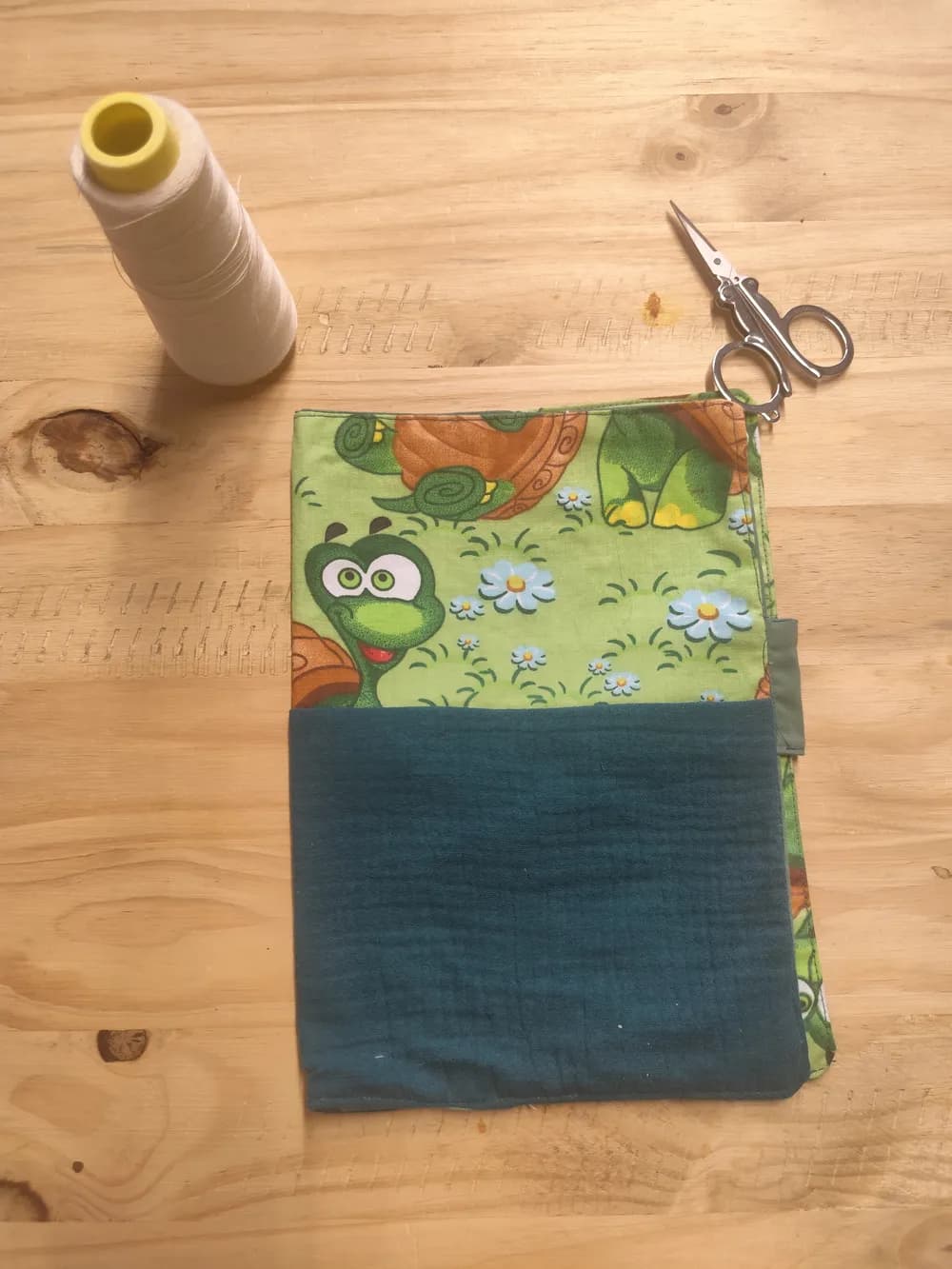 Protège-carnet de santé fermé posé à plat sur une table en bois, partie supérieure en coton vert clair imprimé d'une tortue, d'une grenouille souriante et de petites fleurs blanches sur fond d'herbe, partie inférieure en coton uni vert foncé, languette de fermeture scratch verte visible sur le bord droit, une bobine de fil blanc et une paire de ciseaux posées au-dessus