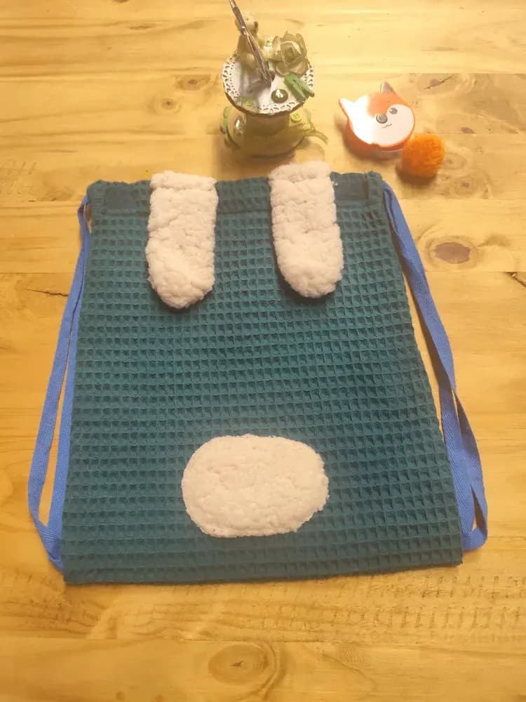 Sac à dos enfant posé bien à plat sur une table en bois clair, vu de face. Le tissu nid d'abeille est bleu canard, avec un fin quadrillage en relief. En haut, deux petites oreilles arrondies en peluche bouclée couleur crème sont cousues, écartées l'une de l'autre. En bas, un grand nez ovale dans la même peluche crème est cousu au centre. Les cordons de fermeture en coton bleu longent les deux côtés du sac. Au-dessus sont posés un pot de fleurs séchées, un petit renard décoratif et un pompon orange. Aucun prénom n'est brodé sur ce modèle photographié