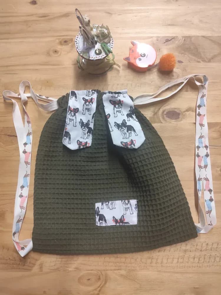 Sac à dos enfant posé à plat sur une table en bois clair, légèrement resserré en haut. Le tissu nid d'abeille est vert kaki, avec un fin quadrillage en relief. En haut, deux oreilles allongées en coton blanc, imprimées de petits bouledogues français noirs et blancs à nœud rouge, pendent côte à côte. En bas, un nez rectangulaire dans le même tissu blanc à bouledogues est cousu au centre. Les bretelles beiges, imprimées de bassets en manteau rose ou bleu, encadrent le sac de chaque côté. Au-dessus sont posés un pot de fleurs séchées, un petit renard décoratif et un pompon orange. Aucun prénom n'est brodé sur ce modèle photographié