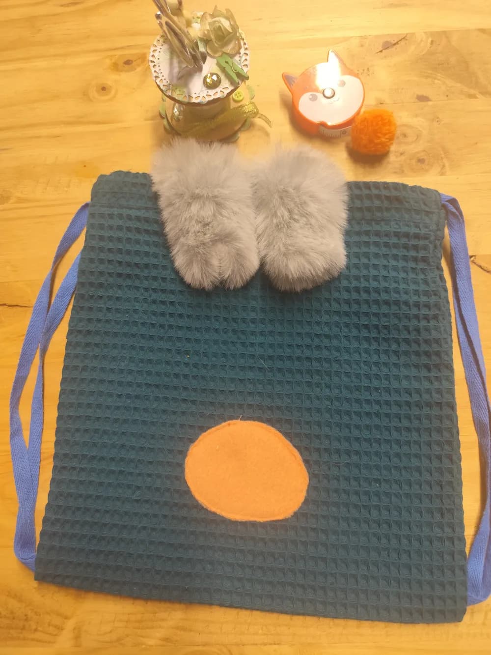 Sac à dos enfant posé à plat sur une table en bois clair, vu de face. Le tissu nid d'abeille est bleu canard foncé, avec un fin quadrillage en relief. En haut, deux oreilles en fausse fourrure grise, douces et épaisses, sont posées côte à côte. En bas, un nez rond en tissu orange est cousu au centre. Les cordons de fermeture en coton bleu longent les deux côtés du sac. Au-dessus sont posés un pot de fleurs séchées, un petit renard décoratif et un pompon orange. Aucun prénom n'est brodé sur ce modèle photographié