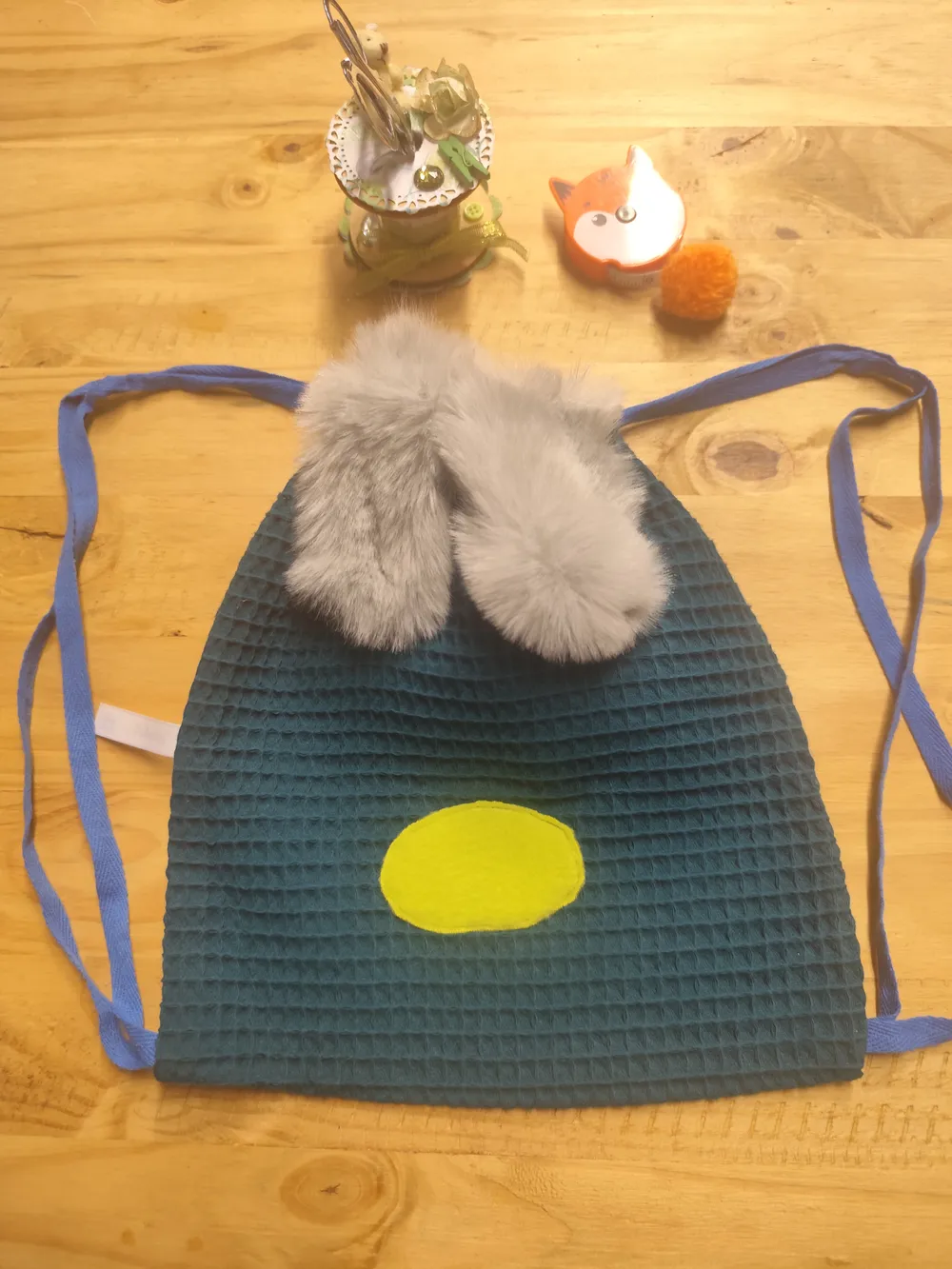 Sac à dos enfant en tissu nid d'abeille bleu canard posé à plat sur une table en bois, deux longues oreilles en fausse fourrure grise retombant en haut du sac, un nez ovale jaune cousu en bas, cordons de fermeture en coton bleu dépliés de chaque côté, un petit renard décoratif, un pompon orange et un pot de fleurs séchées posés au-dessus