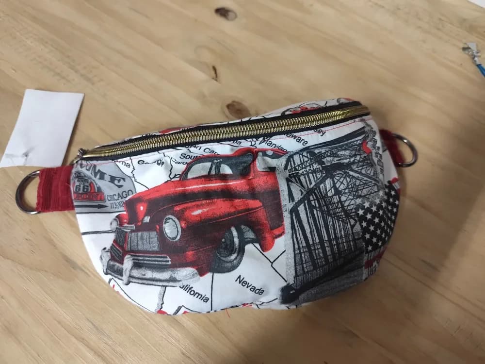 Face avant du sac banane en forme de demi-lune, en tissu blanc imprimé façon route 66 vintage : une grosse voiture américaine rouge des années 40 au centre, un pont métallique en noir et gris à droite, un coin de drapeau américain étoilé, un panneau Route 66 Chicago et des noms d'États comme Nevada et California. Une fermeture éclair dorée court sur tout le haut du sac. De chaque côté, une patte en tissu rouge porte un anneau argenté où s'accroche la sangle. Le sac est posé à plat sur une table en bois clair.