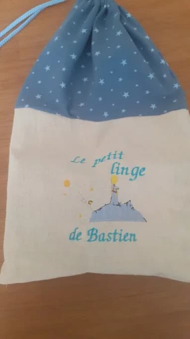 Sac à cordon fait main CÉKIKAFÉ : le haut en coton bleu à petites étoiles blanches, le bas en toile de coton écrue, brodé du Petit Prince sur sa planète et de l'inscription « Le petit linge de Bastien » en fil bleu turquoise. Le cordon bleu clair dépasse en haut à gauche. Posé à plat sur une table en bois.
