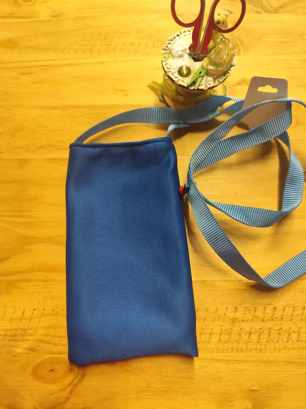 Sac pochette téléphone en tissu bleu uni satiné, longue anse bleu clair, posé sur une table en bois avec son étiquette