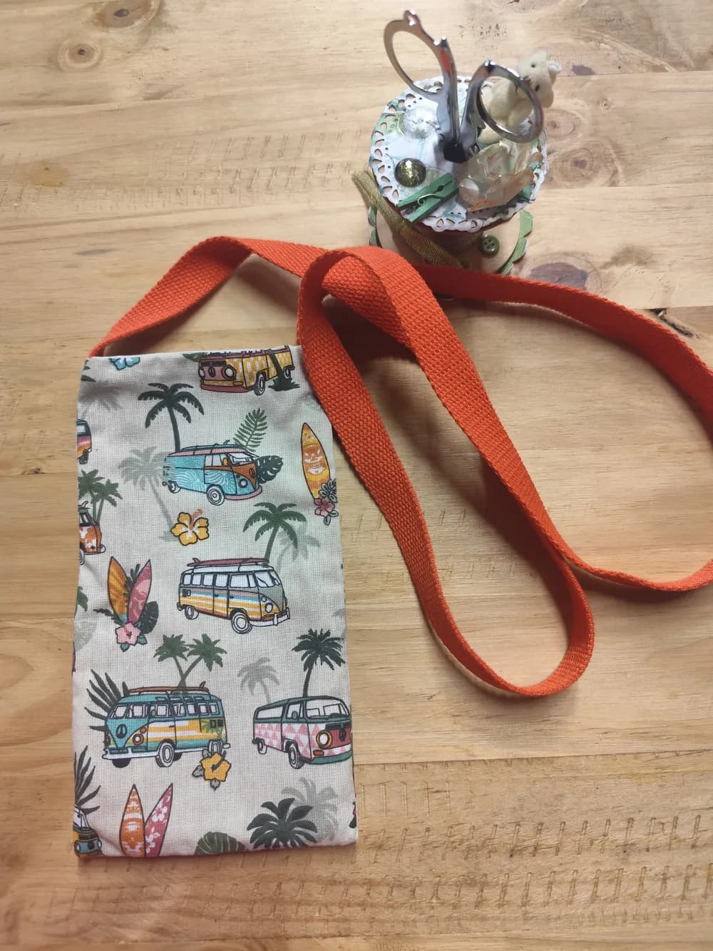 Sac pochette téléphone en tissu beige illustré de vans Volkswagen combi colorés, palmiers et planches de surf, anses en toile orange, posé sur une table en bois