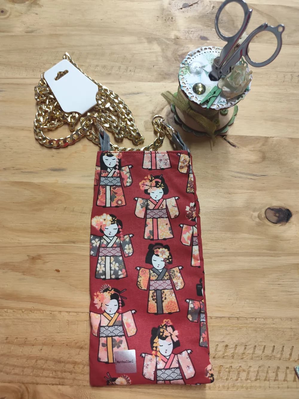 Sac pochette téléphone en tissu rouge à motif de poupées japonaises Kokeshi colorées, chaînette dorée en bandoulière, posé sur une table en bois avec son étiquette