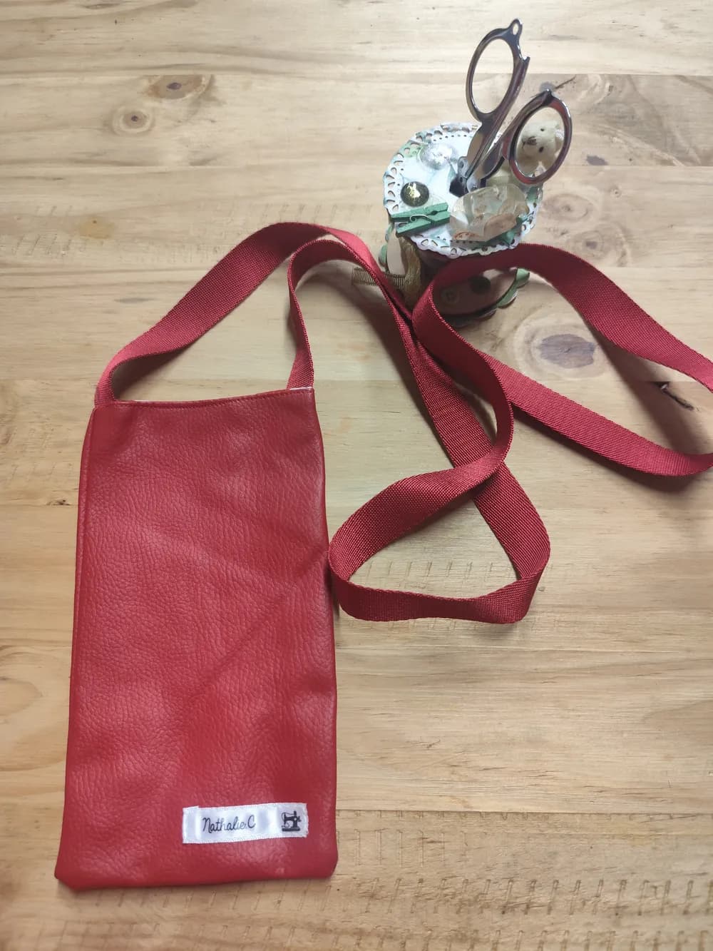Sac pochette téléphone en simili cuir rouge, anses cousues assorties, étiquette « Nathalie C. » cousue en bas, posé sur une table en bois