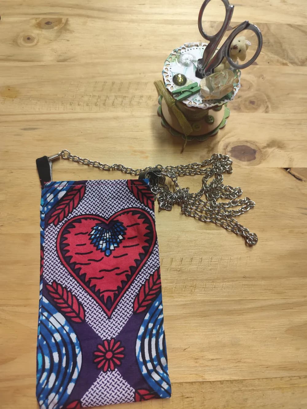 Sac pochette téléphone en tissu africain wax rouge, bleu et blanc à motif cœur central et feuilles, chaîne argentée posée à côté sur une table en bois
