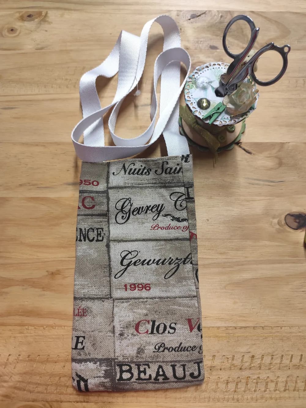 Sac pochette téléphone en tissu beige imprimé d'étiquettes de vin (Nuits-Saint-Georges, Gevrey, Gewurztraminer, Clos Vougeot, Beaujolais), anse large blanche, posé à plat sur une table en bois