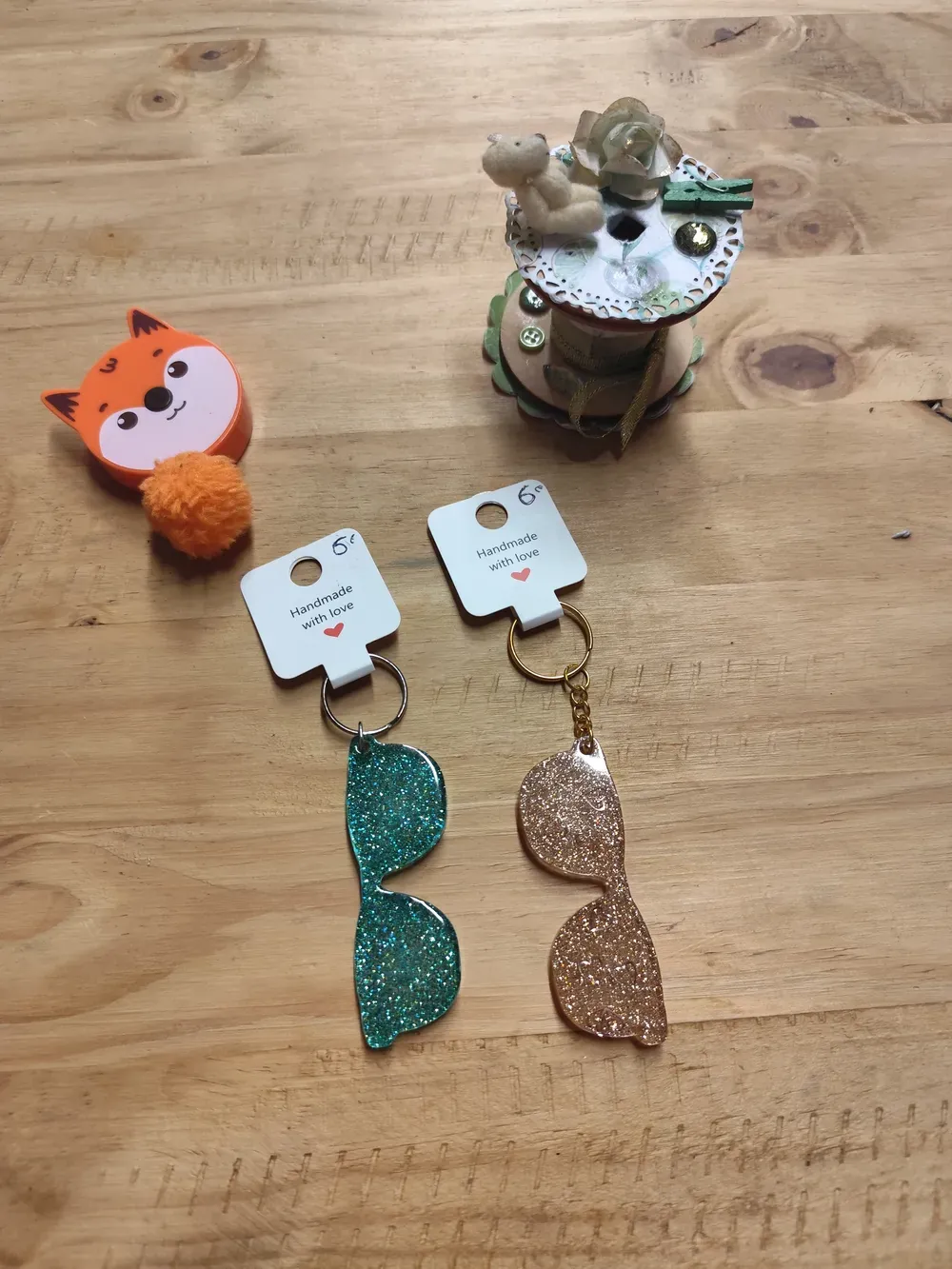 Deux porte-clés CÉKIKAFÉ en forme de lunettes de soleil, en résine époxy pailletée, posés côte à côte sur une table en bois clair, chacun accroché à une étiquette blanche « Handmade with love » avec un cœur rouge et le prix de 6 € écrit à la main. À gauche, une paire vert turquoise couverte de paillettes scintillantes, sur un anneau argenté ; à droite, une paire doré champagne translucide aux fines paillettes dorées, reliée par une petite chaîne à un anneau doré. Autour : un mètre ruban orange en forme de tête de renard avec son pompon orange, et une bobine décorée d'un petit ourson en peluche, de dentelle de papier, d'une fleur et d'une pince à linge verte.