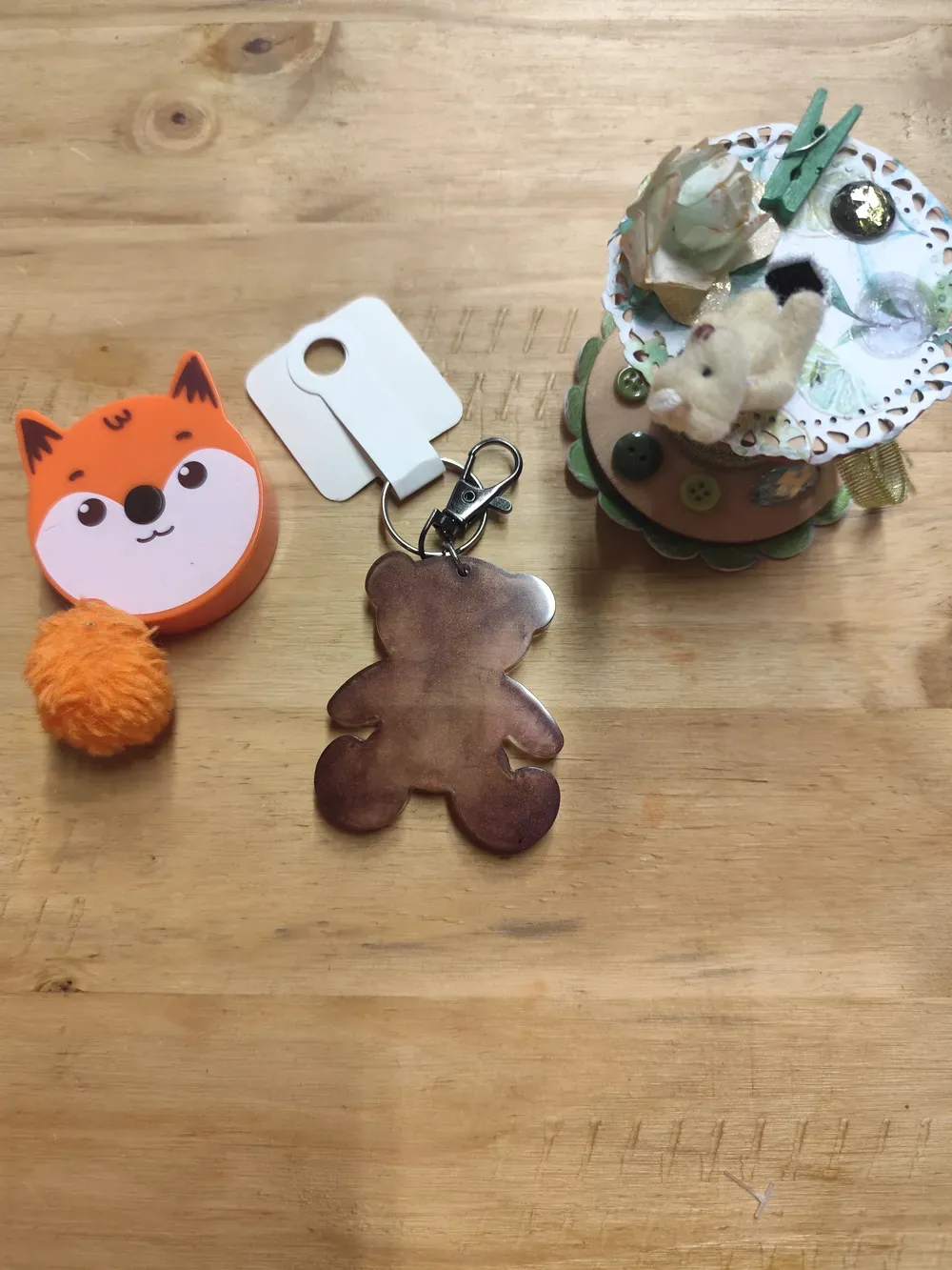 Porte-clés CÉKIKAFÉ en forme d'ourson plat, en résine époxy marron nacré d'une seule couleur, posé à plat sur une table en bois clair : la teinte chocolat au reflet satiné fonce sur les bords et les pieds. Un mousqueton et un anneau argentés le relient à une étiquette cartonnée blanche, vue de dos. À gauche, un mètre ruban orange en forme de tête de renard avec son pompon orange ; à droite, une bobine décorée d'un petit ourson en peluche, de dentelle de papier, d'une fleur, de boutons verts et d'une pince à linge verte.