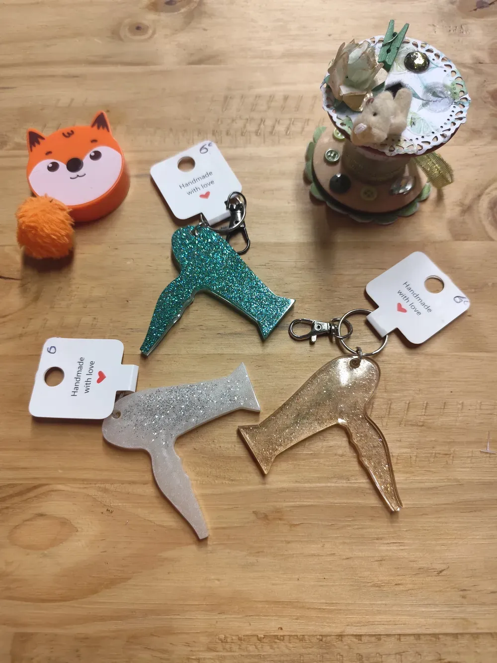 Trois porte-clés CÉKIKAFÉ en forme de sèche-cheveux, en résine époxy pailletée, posés à plat sur une table en bois clair : un vert turquoise couvert de paillettes scintillantes, un blanc nacré parsemé de paillettes argentées et un doré champagne transparent aux fines paillettes dorées. Chacun est accroché par un mousqueton et un anneau argentés à une étiquette blanche « Handmade with love » avec un cœur rouge et le prix de 6 € écrit à la main. Autour : un mètre ruban orange en forme de tête de renard avec son pompon orange, et une bobine décorée d'un petit ourson en peluche, de dentelle de papier, d'une fleur et d'une pince à linge verte.